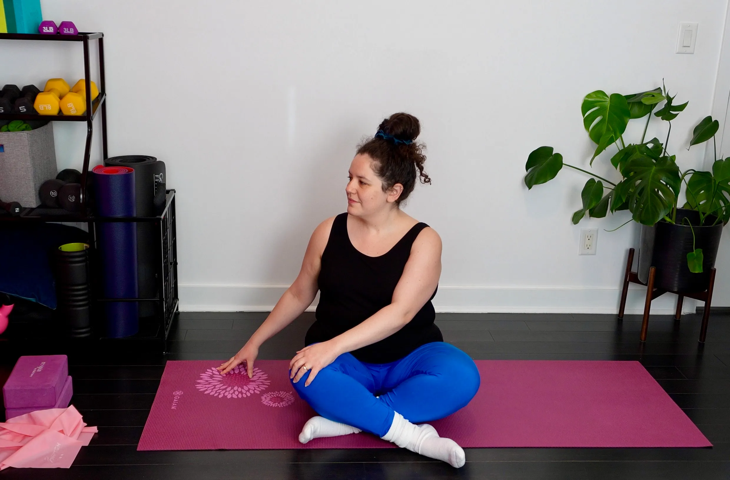 Rebecca Yaffa sitting cross-legged on a pink yoga mat in her home studio, reaching gently toward the floor.