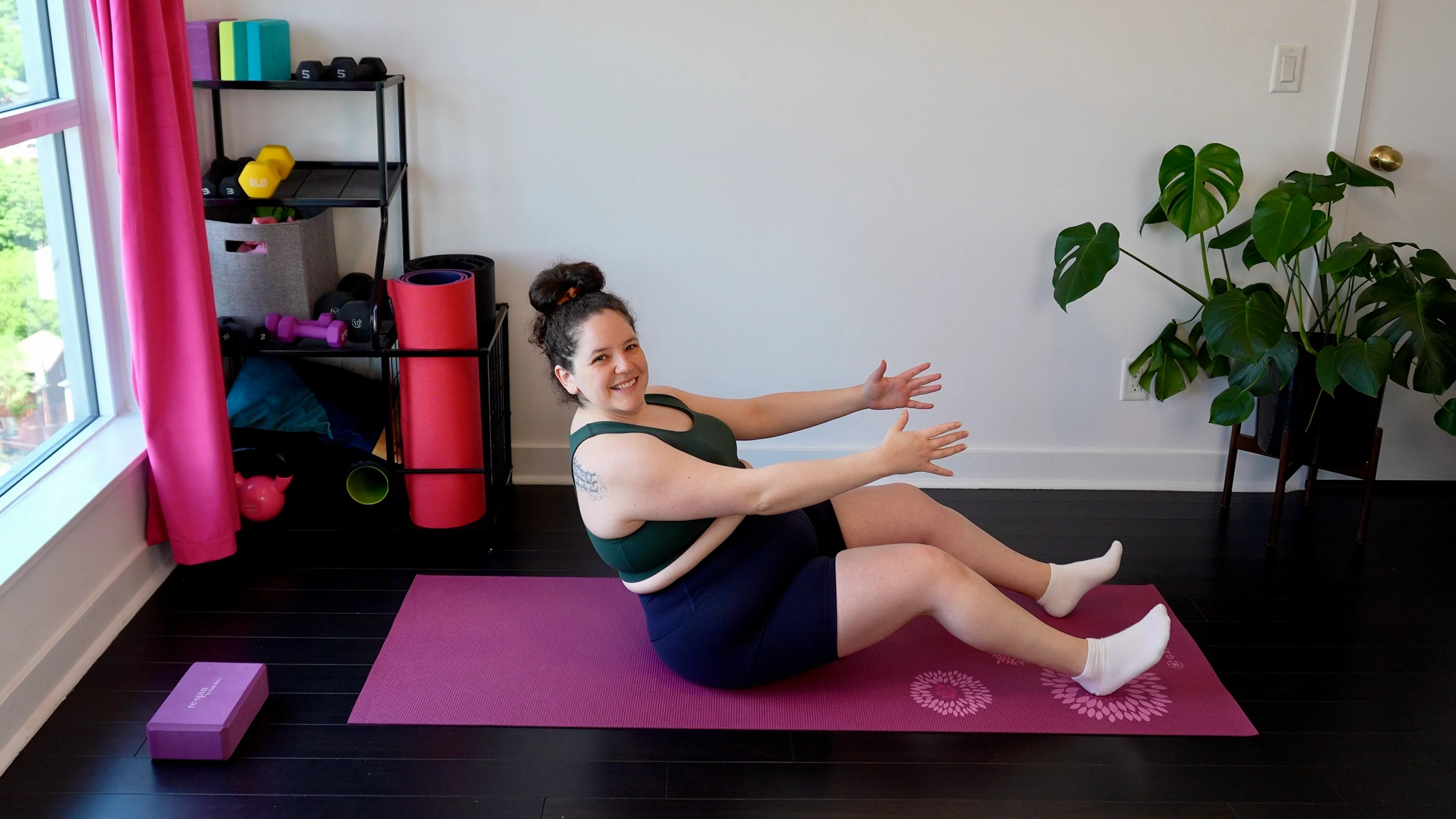 Rebecca Yaffa doing a seated core exercise on a pink yoga mat in her home studio, smiling at the camera.